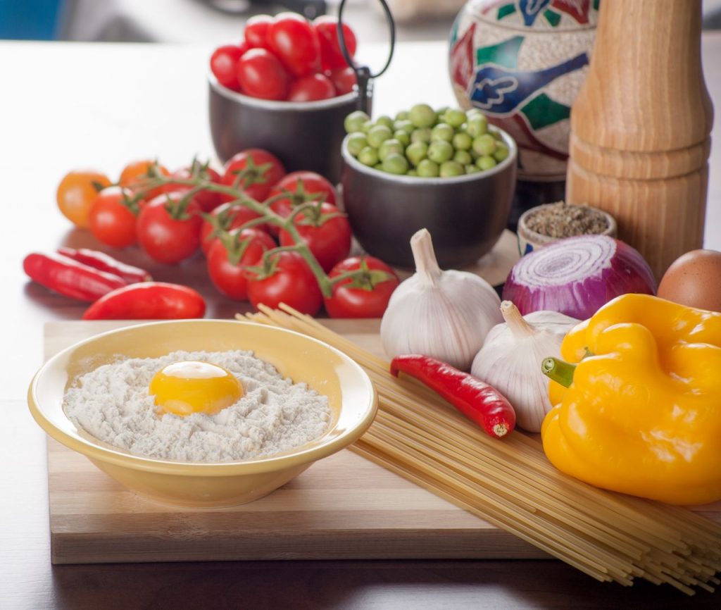 A vibrant assortment of fresh ingredients including vegetables, pasta, and an egg ready for cooking in a cozy kitchen setting.